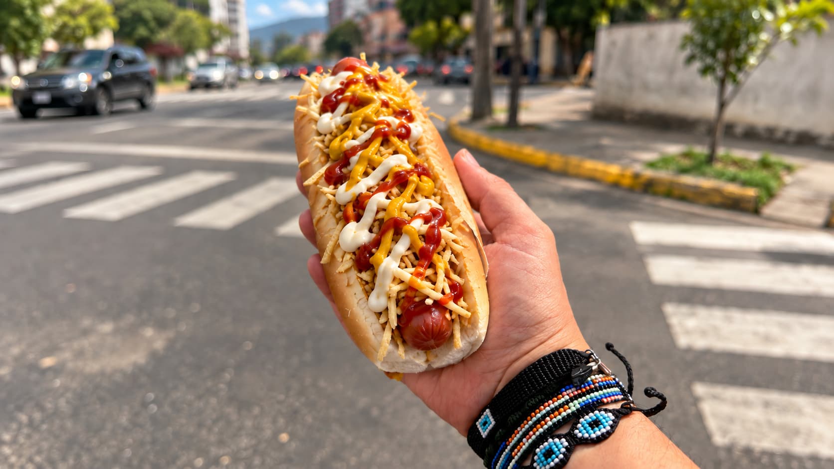 Una mano sostiene un perro caliente con salsas frente a una calle